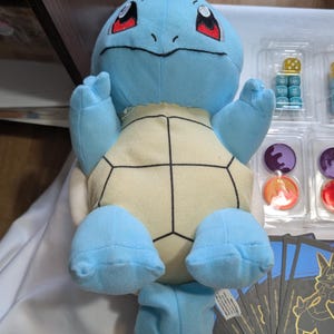May include: A blue and yellow Squirtle plush toy with red eyes and a black outline. The toy is sitting on a table with a set of dice and cards in the background.