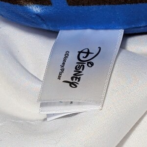 May include: A white tag with the Disney logo and the text "&copy;Disney/Pixar" is visible. The tag is attached to a blue and brown plush toy. The background is a white fabric.