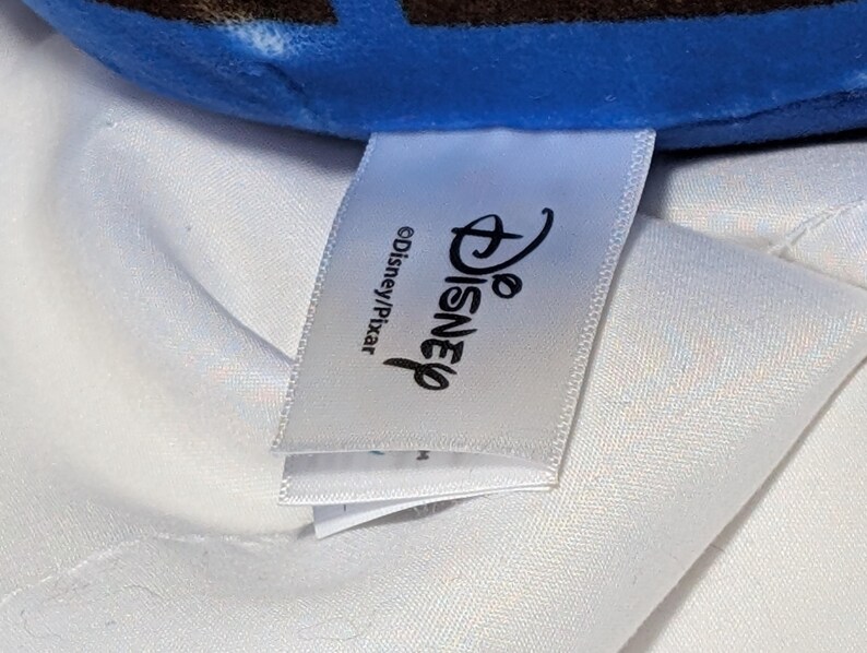 May include: A white tag with the Disney logo and the text "&copy;Disney/Pixar" is visible. The tag is attached to a blue and brown plush toy. The background is a white fabric.