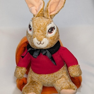 Op de afbeelding: Een pluchen konijn met bruin haar, roze oren en rode ogen. Het konijn draagt een rode jas met een zwarte strik en zit in een oranje wortelvormige draagzak. Het speelgoed is ontworpen om op een personage uit een kinderboek te lijken.
