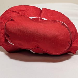 May include: A red, heart-shaped plush cushion with a satin-like texture. The cushion has a white trim and is resting on a white surface. The cushion is a Valentine's Day or romantic-themed item.