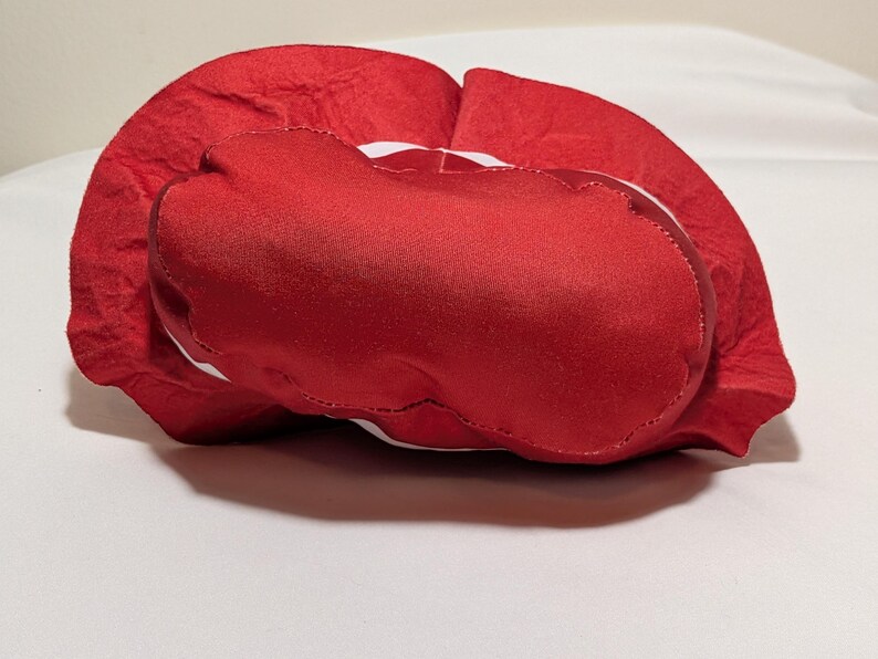 May include: A red, heart-shaped plush cushion with a satin-like texture. The cushion has a white trim and is resting on a white surface. The cushion is a Valentine's Day or romantic-themed item.