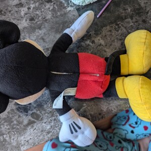 Puede incluir: Un juguete de peluche Mickey Mouse negro y rojo con zapatos amarillos. El juguete est&aacute; acostado de espaldas con los brazos y las piernas extendidos.