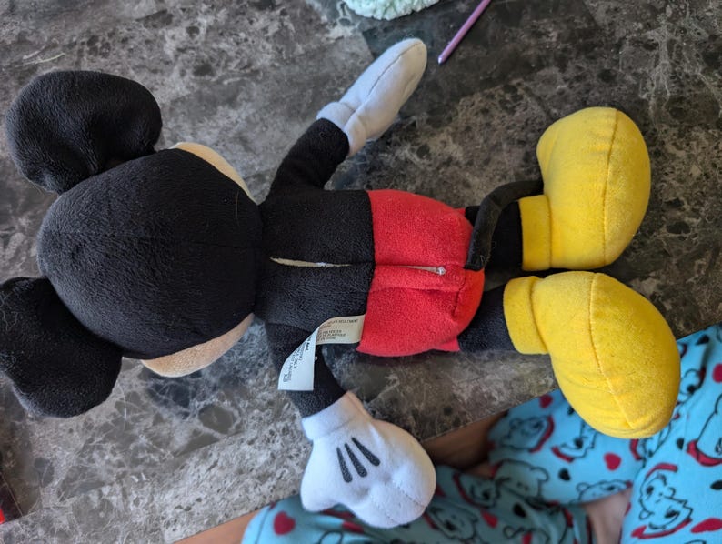 Puede incluir: Un juguete de peluche Mickey Mouse negro y rojo con zapatos amarillos. El juguete est&aacute; acostado de espaldas con los brazos y las piernas extendidos.