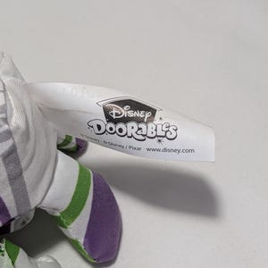 May include: A Disney Doorables plush toy with a white tag featuring the Disney logo and the Doorables brand name. The plush toy has a white, green, and purple color scheme.