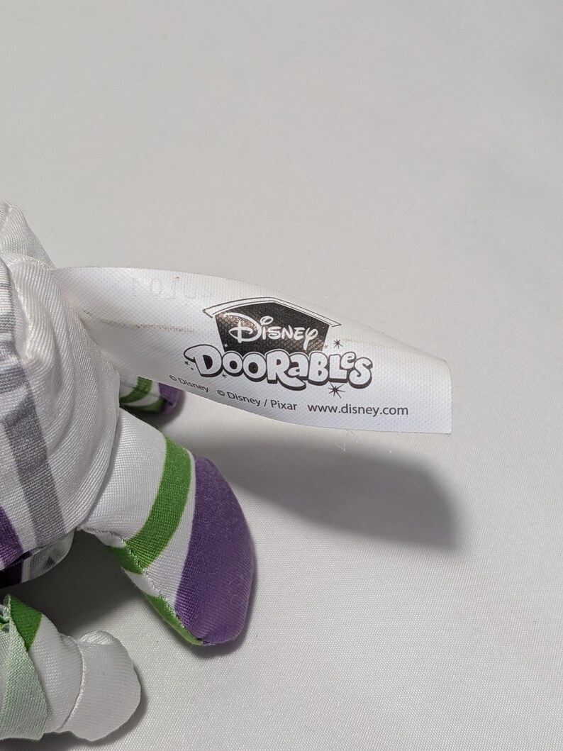May include: A Disney Doorables plush toy with a white tag featuring the Disney logo and the Doorables brand name. The plush toy has a white, green, and purple color scheme.