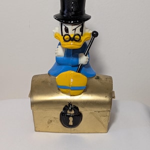 May include: A vintage Scrooge McDuck figurine atop a gold-colored treasure chest. The figurine wears a black top hat, glasses, and a blue coat, holding a cane. The chest has a black lock. A collectible item.