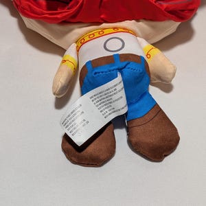 May include: A soft toy of a character with a red hat, tan skin, and a white shirt with a yellow collar. The character wears blue jeans and brown boots. A white tag is attached to the back of the toy.