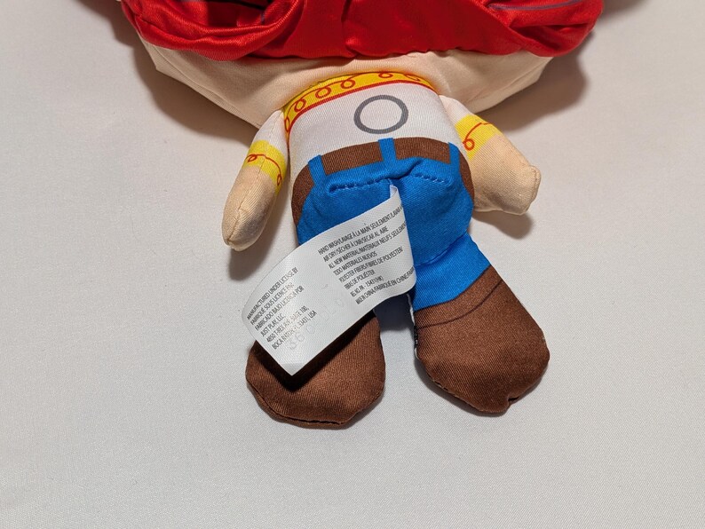 May include: A soft toy of a character with a red hat, tan skin, and a white shirt with a yellow collar. The character wears blue jeans and brown boots. A white tag is attached to the back of the toy.
