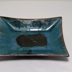 May include: A rectangular ceramic dish with a glossy blue glaze that transitions into dark brown edges. The center features a matte black oval area with cursive gold text written across it. The dish has slightly flared sides and a smooth finish.