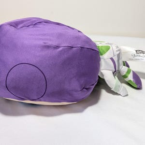 May include: A plush toy of Buzz Lightyear from Disney's Toy Story. The toy features a large purple head with a circular design, and white and green arms. A Disney Doorables tag is attached.
