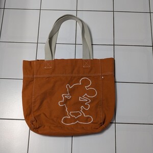 May include: A brown canvas tote bag with white straps and a white embroidered Mickey Mouse silhouette.
