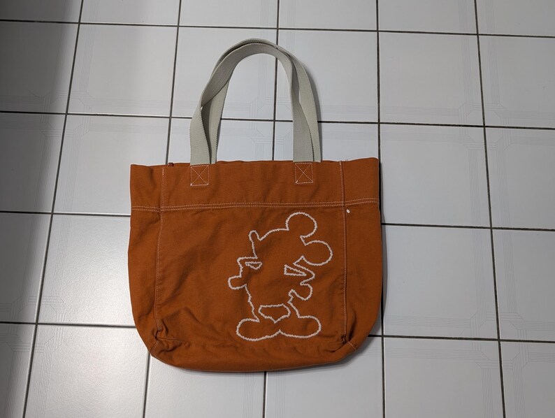 May include: A brown canvas tote bag with white straps and a white embroidered Mickey Mouse silhouette.