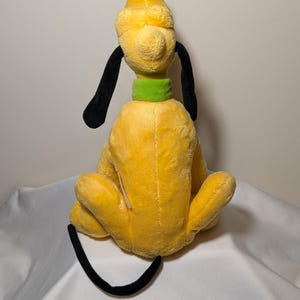 Puede incluir: Un peluche de Pluto, un personaje de perro amarillo con orejas y cola negras, un collar verde y un sombrero amarillo con punta negra. El juguete est&aacute; sentado con las piernas hacia adelante.
