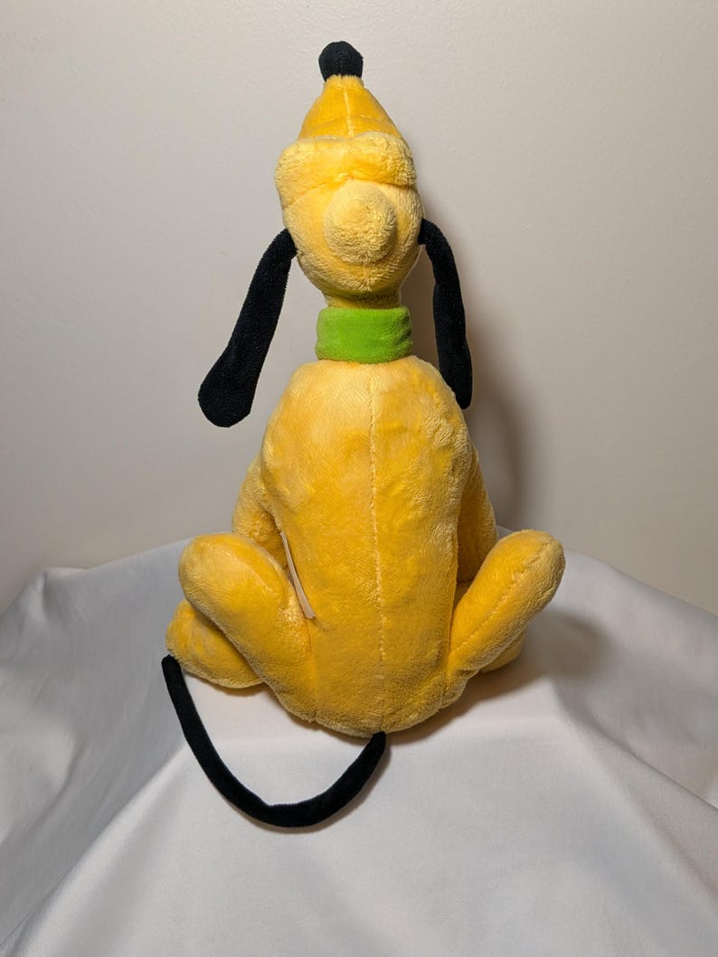 Puede incluir: Un peluche de Pluto, un personaje de perro amarillo con orejas y cola negras, un collar verde y un sombrero amarillo con punta negra. El juguete est&aacute; sentado con las piernas hacia adelante.