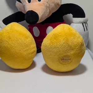 May include: A plush Mickey Mouse toy with yellow feet, red shorts, black arms, and a tan face. The toy has white gloves and a black nose. The bottom of one foot has a Disney Store label.