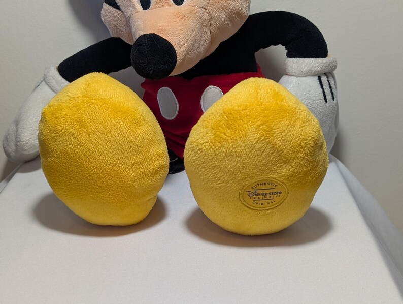 May include: A plush Mickey Mouse toy with yellow feet, red shorts, black arms, and a tan face. The toy has white gloves and a black nose. The bottom of one foot has a Disney Store label.