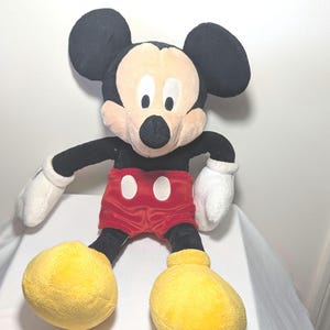 May include: A plush Mickey Mouse toy. The iconic character features large black ears, a cream-colored face, and a black nose. He wears red shorts with white buttons, white gloves, and large yellow shoes. The toy is soft and cuddly.