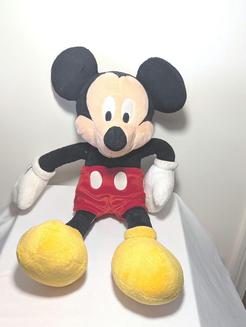 May include: A plush Mickey Mouse toy. The iconic character features large black ears, a cream-colored face, and a black nose. He wears red shorts with white buttons, white gloves, and large yellow shoes. The toy is soft and cuddly.
