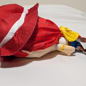 May include: A plush doll of a character wearing a red hat with a white stripe, a red cape, and a yellow bow. The doll has a tan face, blue trousers, and brown boots. The doll is lying on a white surface.