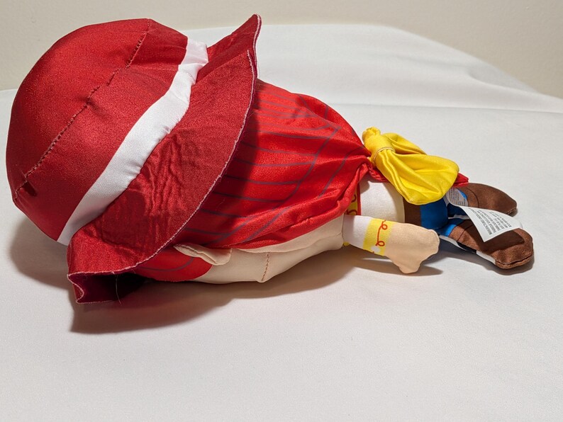 May include: A plush doll of a character wearing a red hat with a white stripe, a red cape, and a yellow bow. The doll has a tan face, blue trousers, and brown boots. The doll is lying on a white surface.