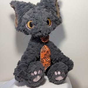 Può includere: Un gatto di peluche grigio con grandi occhi gialli e dettagli rosa. Indossa una cravatta arancione decorata con un motivo a ragnatela nera. Le zampe del gatto presentano cuscinetti rosa. Un peluche a tema Halloween.