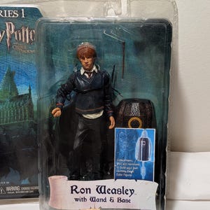 May include: A packaged Ron Weasley action figure from the Harry Potter series. The figure is in a clear plastic case, wearing a dark outfit and holding a wand. The packaging reads "Ron Weasley with Wand & Base."