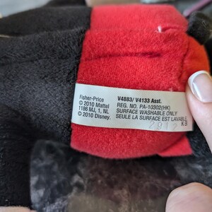 Puede incluir: Un juguete de peluche rojo y negro con una etiqueta blanca que dice "Fisher-Price &copy;2010 Mattel 1186 MJ, 1, NL &copy;2010 Disney. V4883/V4133 Asst. REG. NO. PA-10302(HK) SURFACE WASHABLE ONLY SEULE LA SURFACE EST LAVABLE 2912 KB"
