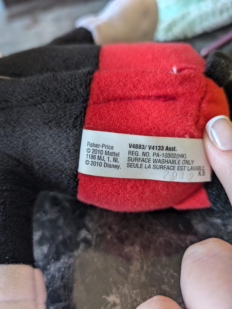 Puede incluir: Un juguete de peluche rojo y negro con una etiqueta blanca que dice "Fisher-Price &copy;2010 Mattel 1186 MJ, 1, NL &copy;2010 Disney. V4883/V4133 Asst. REG. NO. PA-10302(HK) SURFACE WASHABLE ONLY SEULE LA SURFACE EST LAVABLE 2912 KB"