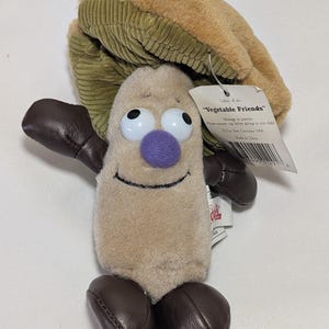 Peut inclure: Une peluche de la collection "Vegetable Friends", représentant un personnage souriant de couleur beige avec un nez violet, de grands yeux et des détails marron. Le jouet a un chapeau en velours côtelé et des mains et des pieds en similicuir marron. Une étiquette indique "Vegetable Friends".