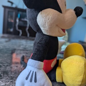 Puede incluir: Un juguete de peluche de Mickey Mouse con pelaje negro, pantalones cortos rojos y zapatos amarillos. El juguete est&aacute; sentado con la mano derecha extendida y la mano izquierda descansando en su regazo.