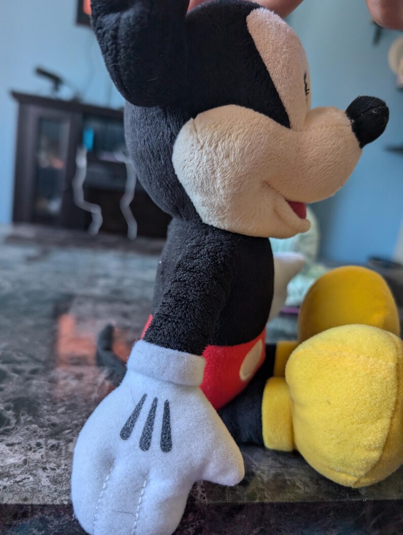 Puede incluir: Un juguete de peluche de Mickey Mouse con pelaje negro, pantalones cortos rojos y zapatos amarillos. El juguete est&aacute; sentado con la mano derecha extendida y la mano izquierda descansando en su regazo.