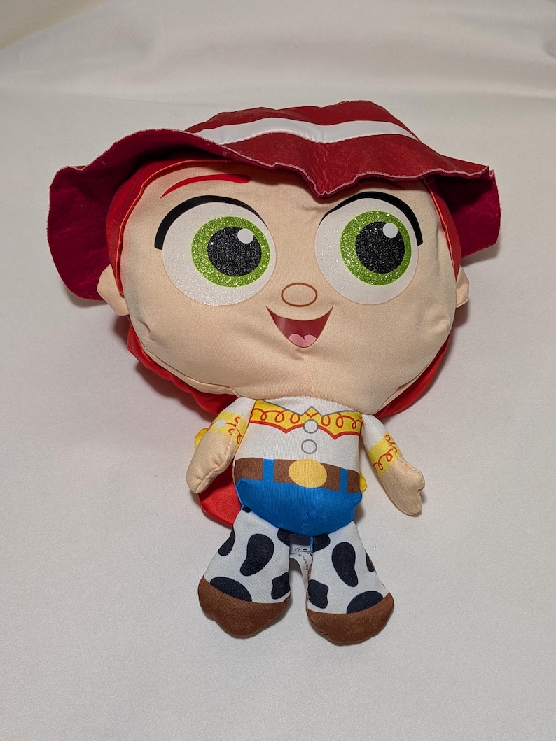 May include: A plush doll of a character with red hair and a matching hat. The doll has large green eyes, a tan face, and a white shirt with a yellow collar. The doll's blue trousers have black and white spots. The doll is a character from a popular animated film.