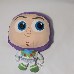 May include: A plush toy of the character Buzz Lightyear from the Toy Story franchise. The toy features a large, round head with a purple hood, large blue eyes with glitter, and a small smile. The body is white with green and blue accents.