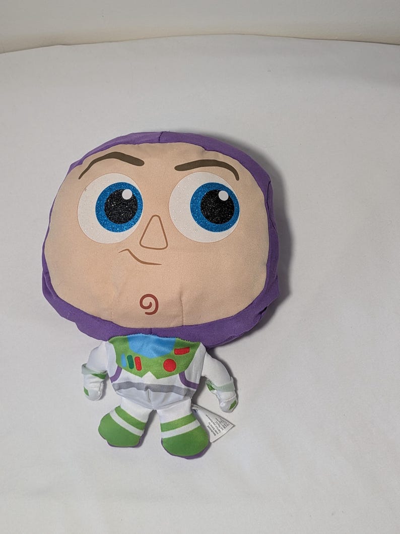 May include: A plush toy of the character Buzz Lightyear from the Toy Story franchise. The toy features a large, round head with a purple hood, large blue eyes with glitter, and a small smile. The body is white with green and blue accents.