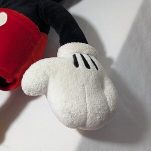 May include: Close-up of a plush Mickey Mouse toy's white glove with three black lines on the palm. The glove is soft and fluffy, with a rounded shape. The toy's red shorts and black arm are also visible.
