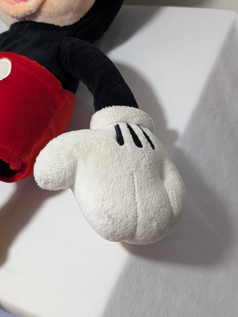 May include: Close-up of a plush Mickey Mouse toy's white glove with three black lines on the palm. The glove is soft and fluffy, with a rounded shape. The toy's red shorts and black arm are also visible.