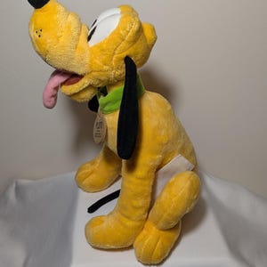 Puede incluir: Un peluche amarillo de Pluto con orejas negras, nariz negra y lengua rosa. El juguete tiene un collar verde y una etiqueta que dice "Pluto". El juguete est&aacute; sobre una superficie blanca.