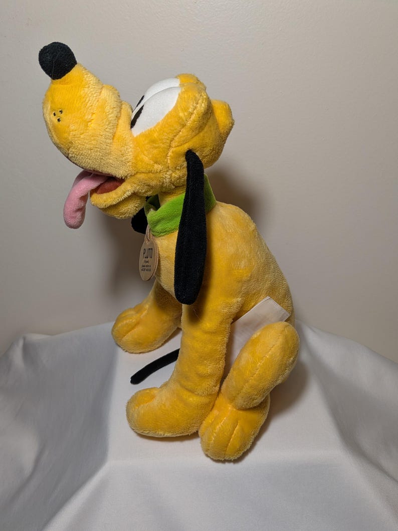 Puede incluir: Un peluche amarillo de Pluto con orejas negras, nariz negra y lengua rosa. El juguete tiene un collar verde y una etiqueta que dice "Pluto". El juguete est&aacute; sobre una superficie blanca.