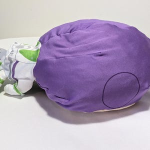 May include: A plush toy in the shape of a character from a children's animated film. The toy is primarily purple with white, green, and gray accents. The toy is lying on a white surface.
