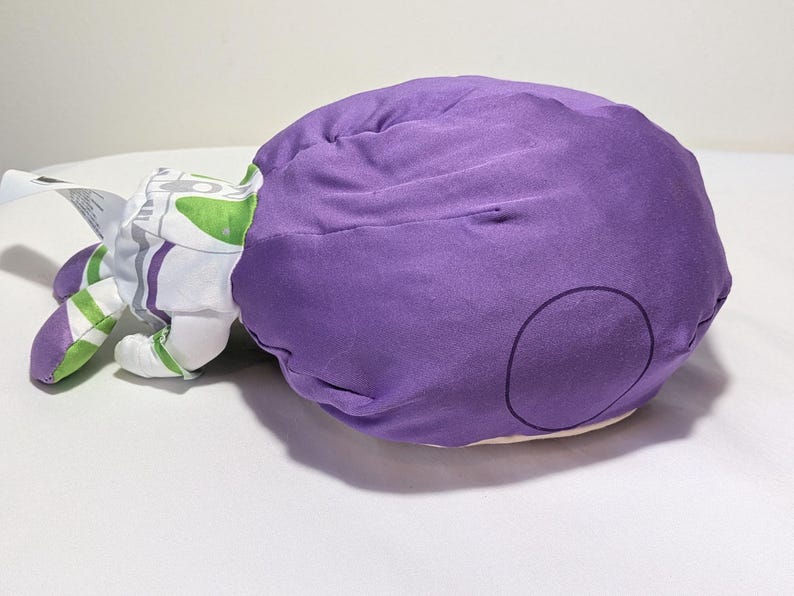 May include: A plush toy in the shape of a character from a children's animated film. The toy is primarily purple with white, green, and gray accents. The toy is lying on a white surface.