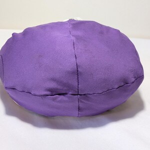 May include: A round, purple fabric object, possibly a pillow or cushion, is centered in the image. The object is made of a solid, textured material and is stitched together with visible seams. The background is a plain white surface.