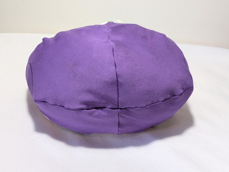 May include: A round, purple fabric object, possibly a pillow or cushion, is centered in the image. The object is made of a solid, textured material and is stitched together with visible seams. The background is a plain white surface.