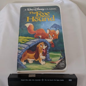 May include: A VHS tape of the Disney animated classic, "The Fox and the Hound." The cover art features a red fox and a brown hound dog. The VHS tape is black and has text that reads "Insert this side into recorder."