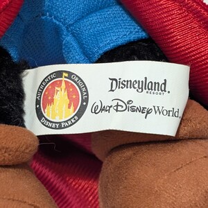 May include: Close-up of a plush toy with a red and blue outfit. The toy has brown arms and a black head. A white tag reads "Disneyland Resort" and "Walt Disney World."