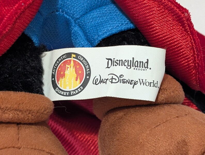 May include: Close-up of a plush toy with a red and blue outfit. The toy has brown arms and a black head. A white tag reads "Disneyland Resort" and "Walt Disney World."