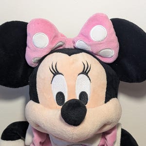 May include: A plush Minnie Mouse doll with large black ears and a pink bow with white polka dots. The doll has a light pink face, black eyes, and a black nose. The doll is wearing a pink outfit.