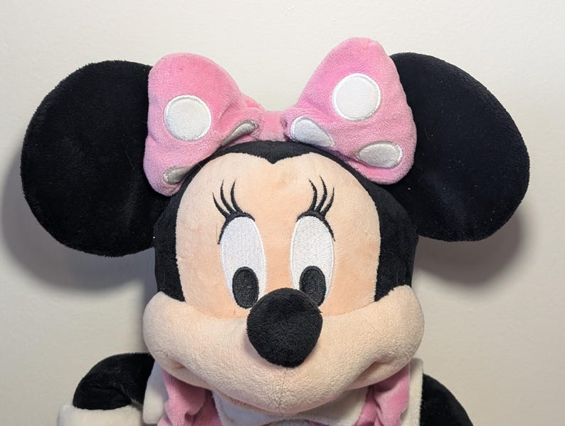 May include: A plush Minnie Mouse doll with large black ears and a pink bow with white polka dots. The doll has a light pink face, black eyes, and a black nose. The doll is wearing a pink outfit.