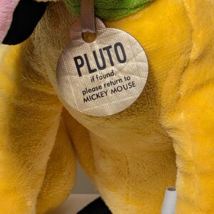 Puede incluir: Un peluche amarillo de Pluto con un collar verde y una etiqueta que dice "PLUTO, if found, please return to MICKEY MOUSE". El juguete tiene detalles en rosa y negro.