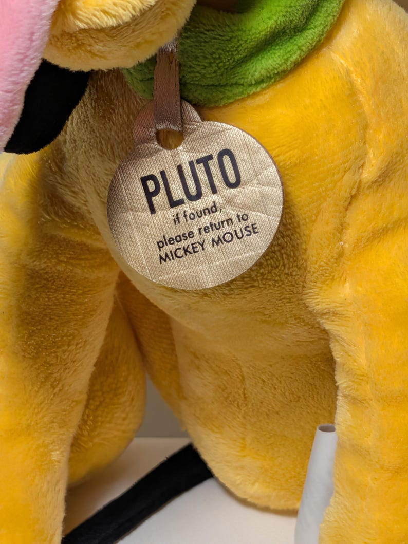 Puede incluir: Un peluche amarillo de Pluto con un collar verde y una etiqueta que dice "PLUTO, if found, please return to MICKEY MOUSE". El juguete tiene detalles en rosa y negro.